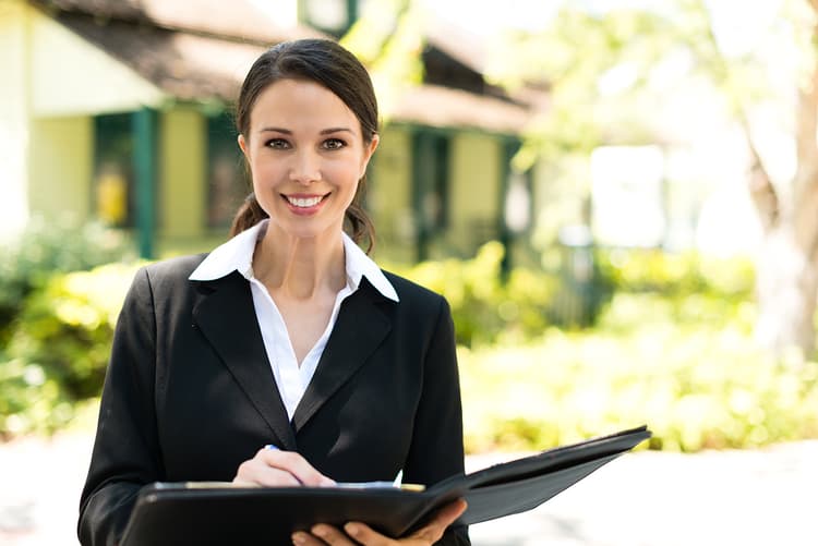 Woman wearing a suit and holding a portfolio with a house in the background